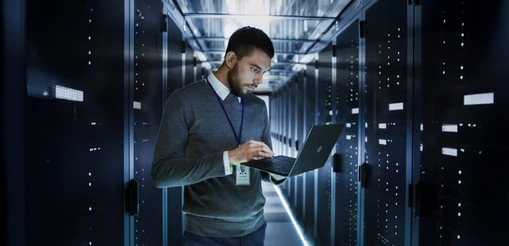IT Technician Works on a Laptop in Big Data Center full of Rack Servers. He Runs Diagnostics and Maintenance, Sets up System.